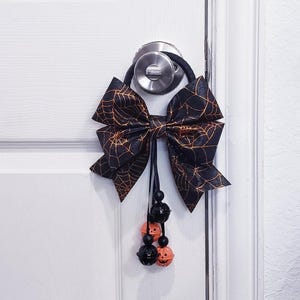 Pode incluir: Um laço de porta de Halloween preto e laranja com um design de teia de aranha. O laço é preso a uma faixa elástica preta e pendurado em uma maçaneta. Contas pretas e enfeites em forma de abóbora laranja pendem do laço.