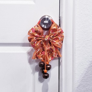 Pode incluir: Um pendente de porta em tecido com um laço grande com um padrão de outono de cogumelos e folhas em tons de laranja, vermelho e castanho. O laço é acentuado com fitas cor de laranja e três sinos metálicos escuros suspensos por contas de madeira.