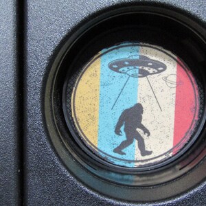 May include: A round coaster with a colorful design featuring a yellow, blue, and red striped background. A black silhouette of a Bigfoot figure is walking towards a flying saucer with a blue and white design. The saucer has a ring of lights around it and is emitting a beam of light.