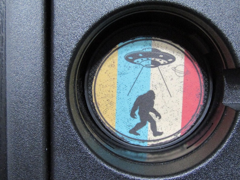 May include: A round coaster with a colorful design featuring a yellow, blue, and red striped background. A black silhouette of a Bigfoot figure is walking towards a flying saucer with a blue and white design. The saucer has a ring of lights around it and is emitting a beam of light.