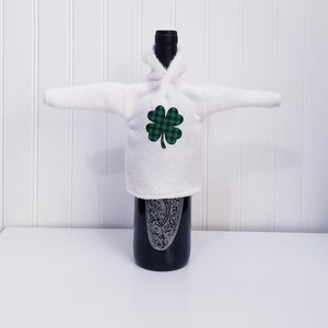 Peut inclure: Une housse de bouteille de vin blanche en forme de sweat à capuche, ornée d'un trèfle à carreaux vert et noir. La bouteille sombre présente un motif orné. Le fond est blanc.