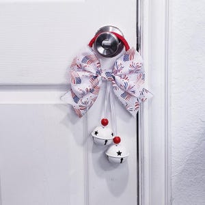 Puede incluir: Adorno patriótico para puerta con un lazo grande con estampado de banderas americanas y fuegos artificiales. Dos campanas blancas con recortes de estrellas y detalles rojos cuelgan debajo. El lazo está sujeto a un lazo rojo.