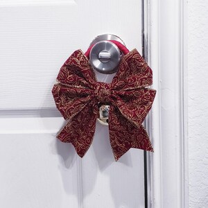 Op de afbeelding: Een decoratieve bordeauxrode strik met gouden accenten hangt aan een witte deur. De strik is gemaakt van stof met een gedetailleerd bloemmotief. Een rood koord is om de deurklink gewikkeld.