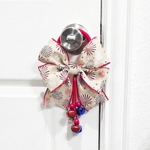 May include: A red, white, and blue bow with a firework pattern and a red tassel with bells hanging from it. The bow is tied around a door knob.