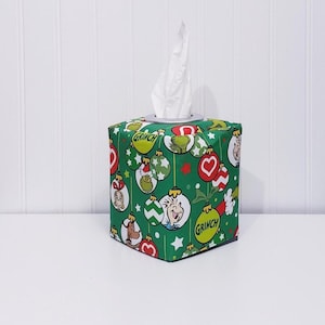 May include: A square tissue box cover with a festive Grinch-themed design. The green fabric is adorned with Grinch characters and ornaments. White tissues are visible at the top. The box is on a white surface.