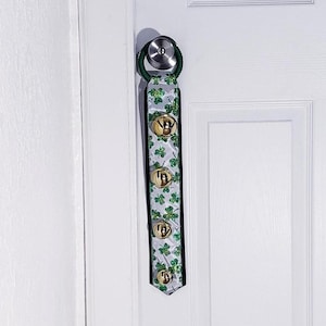 May include: A white door with a green and gold shamrock door knocker with bells. The door knocker is attached to the door knob.