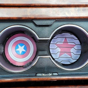 May include: Two car cup holder coasters. One coaster features Captain America's shield design in red, white, and blue. The other coaster has a red star design on a gray and white background.