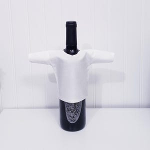 May include: A black wine bottle with a white fabric shirt wrapped around it. The bottle has a decorative label. The background is a white wall and a white surface.