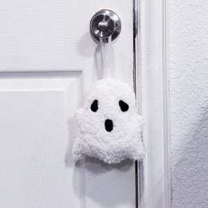 May include: A white plush ghost decoration with a fuzzy, textured surface and black felt eyes and an oval mouth. The ghost is suspended by a thin white ribbon from a silver metal door handle attached to a white paneled door.