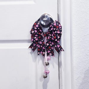 Könnte beinhalten: Eine große dekorative Schleife mit schwarzem Hintergrund und einem Muster aus rosa und roten Herzen. Die Schleife hängt an einer Tür und hat rosa Bänder mit drei kleinen silbernen Glöckchen.