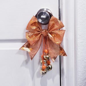 May include: A decorative door bow in shades of orange and gold, attached to a silver doorknob. The bow is made of a sheer, textured fabric. Dangling from the bow are small gold bells and orange beads, adding a festive touch to the door.
