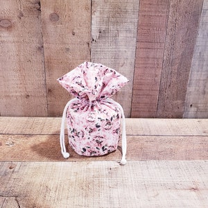 K&ouml;nnte beinhalten: Ein Kordelzugbeutel mit einem floralen Muster in Rosa- und Wei&szlig;t&ouml;nen. Der Beutel ist mit einer wei&szlig;en Kordel gebunden. Der Hintergrund ist eine Holzoberfl&auml;che.