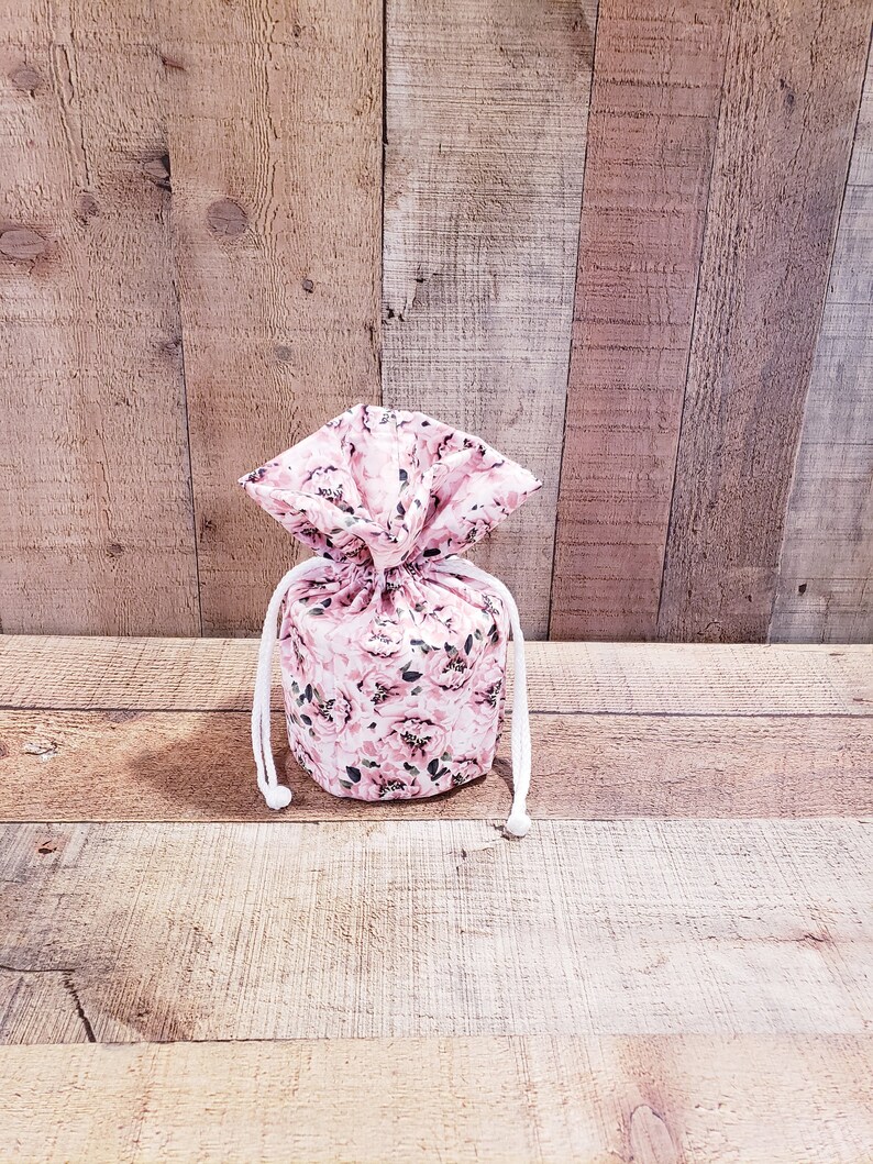 K&ouml;nnte beinhalten: Ein Kordelzugbeutel mit einem floralen Muster in Rosa- und Wei&szlig;t&ouml;nen. Der Beutel ist mit einer wei&szlig;en Kordel gebunden. Der Hintergrund ist eine Holzoberfl&auml;che.
