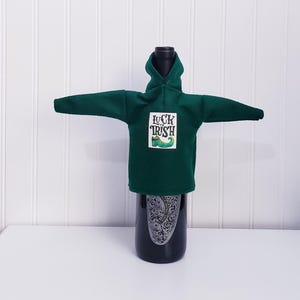 Könnte beinhalten: Dunkelgrüne Weinflaschenhülle in Form eines Hoodies, mit dem Schriftzug "Luck of the Irish" und einem grünen Kleeblatt-Design. Die Hülle befindet sich auf einer schwarzen Weinflasche.