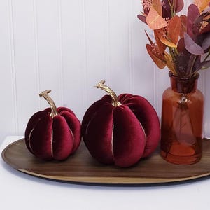 May include: Two plush, burgundy velvet pumpkins with gold stems sit on a wooden tray. A brown glass vase with autumn foliage is to the right. The pumpkins have white stitching and are a seasonal decoration.