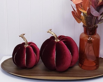 May include: Two plush, burgundy velvet pumpkins with gold stems sit on a wooden tray. A brown glass vase with fall foliage is to the right. The pumpkins have white stitching and are a seasonal decoration.