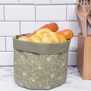 Puede incluir: Una cesta de pan redonda de tela verde oliva llena de panecillos y hogazas doradas. La cesta tiene un estampado floral y un borde verde liso. Cucharas de madera en un soporte de madera a la derecha, y azulejos blancos en el fondo.