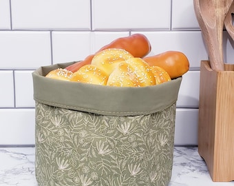 Op de afbeelding: Een ronde broodmand van olijfgroene stof gevuld met goudbruine broodjes en broden. De mand heeft een bloemenpatroon en een effen groene rand. Houten lepels staan in een houten houder rechts en witte tegels op de achtergrond.