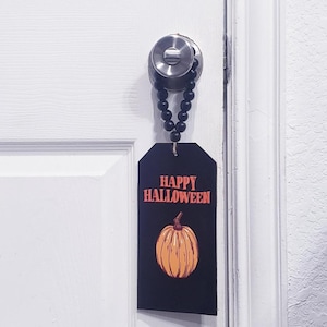 Pode incluir: Uma placa retangular preta para porta com o texto laranja HAPPY HALLOWEEN e uma ilustração de uma abóbora laranja. O objeto está suspenso numa maçaneta prateada por um fio de contas de madeira preta mate preso a um laço de juta.