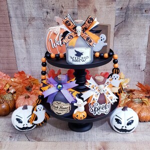 Könnte beinhalten: Ein zweistöckiges schwarzes Tablett mit Halloween-Dekorationen. Es zeigt dekorative Flaschen mit den Aufschriften "Witch's Brew" und "Spooky", Kürbisse, Geisterfiguren und Perlenkränze. Das Farbschema umfasst Orange, Schwarz und Weiß.