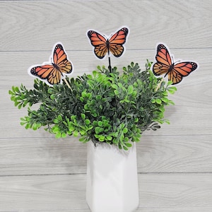 May include: A potted plant with three orange and black butterfly decorations. The butterflies are attached to sticks and are sticking out of the plant.