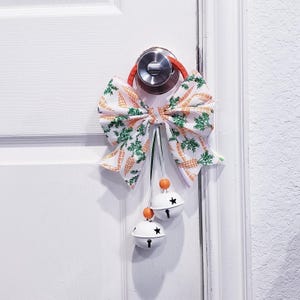 May include: A decorative door bow with a white fabric printed with carrots and green leaves. The bow is attached to a door handle with an orange loop. Two white bells with star cutouts and orange beads hang below.