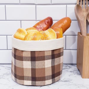 May include: A brown plaid fabric basket filled with various types of bread. The basket has a cream-colored interior and is sitting on a marble countertop. Wooden utensils are in a wooden holder in the background.
