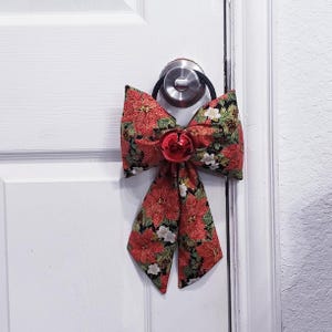 Könnte beinhalten: Eine dekorative Stoffschleife mit einem Muster aus roten Weihnachtssternen, grünen Blättern und kleinen weißen Blüten auf schwarzem Grund. Die Schleife ist mit einer glänzenden roten Glocke versehen und an einem schwarzen Gummiband befestigt.