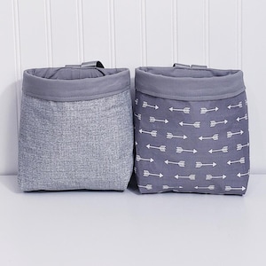 May include: Two grey fabric storage bins. One has a textured pattern, the other features a pattern of white arrows on a grey background. Both have a grey rolled top and a small fabric loop.