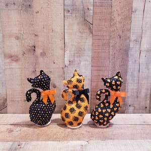 May include: Three black fabric Halloween cat figurines with orange bows. The cats are standing on a wooden surface. The cat on the left has black and white polka dots, the cat in the middle has a black cat print, and the cat on the right has a candy corn print.