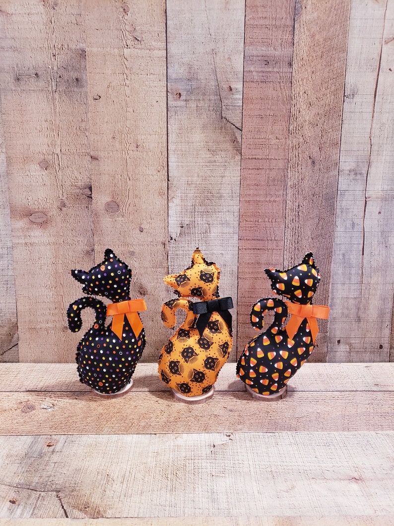 May include: Three black fabric Halloween cat figurines with orange bows. The cats are standing on a wooden surface. The cat on the left has black and white polka dots, the cat in the middle has a black cat print, and the cat on the right has a candy corn print.