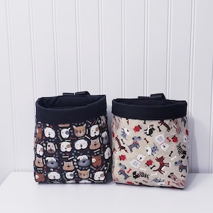 May include: Two black fabric dog treat pouches with a black trim. One pouch has a black background with a white, brown, and red dog print. The other pouch has a beige background with a white, brown, and red dog print.
