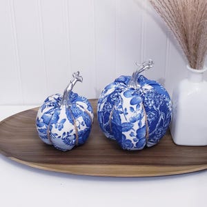 May include: Two fabric pumpkins featuring a blue and white floral pattern. Each pumpkin is topped with a metallic silver stem and is segmented by thin, light brown twine. The fabric displays intricate botanical illustrations in shades of blue.