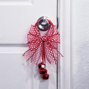 Puede incluir: Un colgador de puerta con lazo rojo y blanco con estampado de corazones y tres campanas rojas colgando debajo. El lazo está sujeto a un anillo rojo que se ajusta a un pomo de puerta plateado. La puerta es blanca.