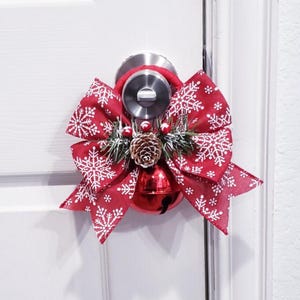 May include: A Christmas door decoration featuring a red bow with white snowflakes, a pine cone, berries, and a jingle bell. The decoration is attached to a door handle, ideal for festive seasonal decor.