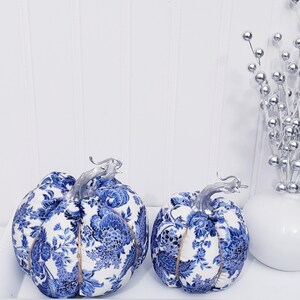 May include: Two decorative fabric pumpkins with blue and white floral patterns. The pumpkins have silver stems and are displayed on a white tray. A vase with silver decorative stems is also present.