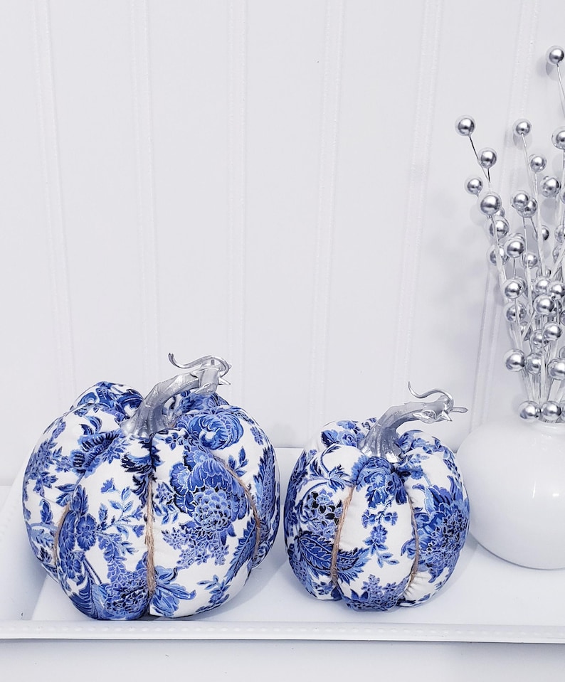 May include: Two decorative fabric pumpkins with blue and white floral patterns. The pumpkins have silver stems and are displayed on a white tray. A vase with silver decorative stems is also present.
