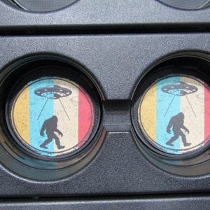 May include: Two round drink coasters with a retro design featuring a Bigfoot walking under a flying saucer. The coasters have a yellow, blue, red, and white color scheme.