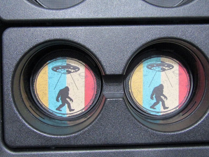 May include: Two round drink coasters with a retro design featuring a Bigfoot walking under a flying saucer. The coasters have a yellow, blue, red, and white color scheme.