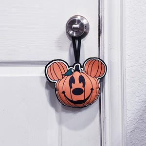 May include: A plush hanging decoration shaped like a pumpkin with mouse ears, featuring a black stitched face, a small green leaf on top, and a black ribbon loop for hanging. The orange pumpkin has black outlines and a smiling expression.