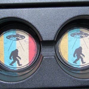 May include: Two round car coasters with a retro design featuring a Bigfoot walking under a UFO. The coasters have a yellow, blue, and red striped background.