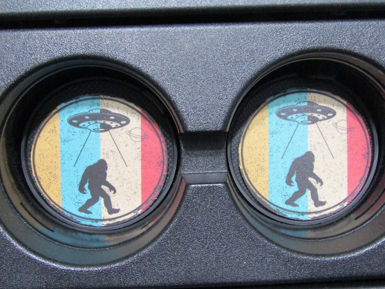 May include: Two round car coasters with a retro design featuring a Bigfoot walking under a UFO. The coasters have a yellow, blue, and red striped background.