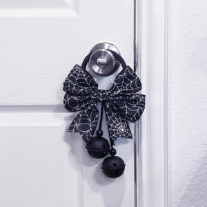 May include: A black and white spiderweb-patterned bow door hanger with two black bells. The bow is attached to a black loop that hangs over a silver doorknob. The bells have a star cut-out design.