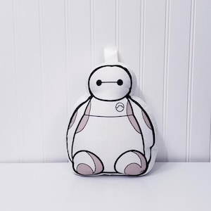 May include: White plush toy of Baymax from the Disney movie Big Hero 6. The toy is sitting with its arms at its sides and has a black outline and pink accents.