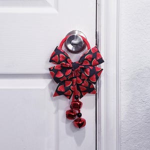 May include: A door hanger with a watermelon-patterned bow on a black background, attached to a red loop. It features three red bells and black beads. The bow is hanging on a white door. The bow's design includes red watermelon slices.