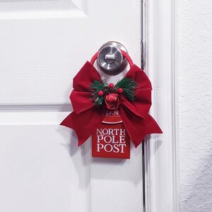 May include: A red door hanger featuring a large fabric bow, green pine needles, small pinecones, and red berries. The rectangular sign has the text NORTH POLE POST in white lettering. The hanger is suspended from a silver-colored metal door knob.