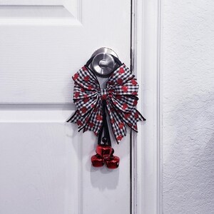 Puede incluir: Adorno para puerta con un gran lazo a cuadros blancos y negros, adornado con detalles de corazones rojos. Tres campanas rojas cuelgan debajo del lazo, suspendidas de un lazo negro. La puerta es blanca.