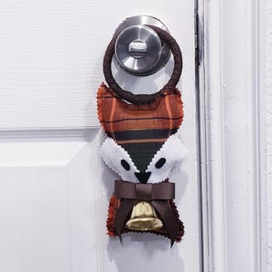 Puede incluir: Un colgador de puerta artesanal con forma de zorro con un patrón de cuadros en tonos naranja, marrón y negro. El zorro tiene una cara blanca, un lazo marrón y una pequeña campana dorada. El colgador está sujeto a un lazo de tela marrón.