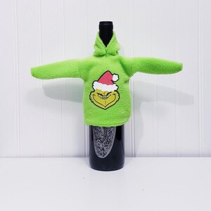 May include: A green bottle cover shaped like a hoodie, featuring the Grinch's face wearing a Santa hat. The cover is made of a fuzzy material and is designed to fit over a wine bottle. The bottle is black with a decorative label.