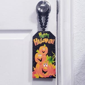 Pode incluir: Uma placa retangular suspensa com fundo preto apresentando três abóboras laranja empilhadas com rostos sorridentes e folhas verdes. O texto "HAPPY HALLOWEEN!" está impresso em laranja e verde no topo. A placa pende de um laço de contas pretas.